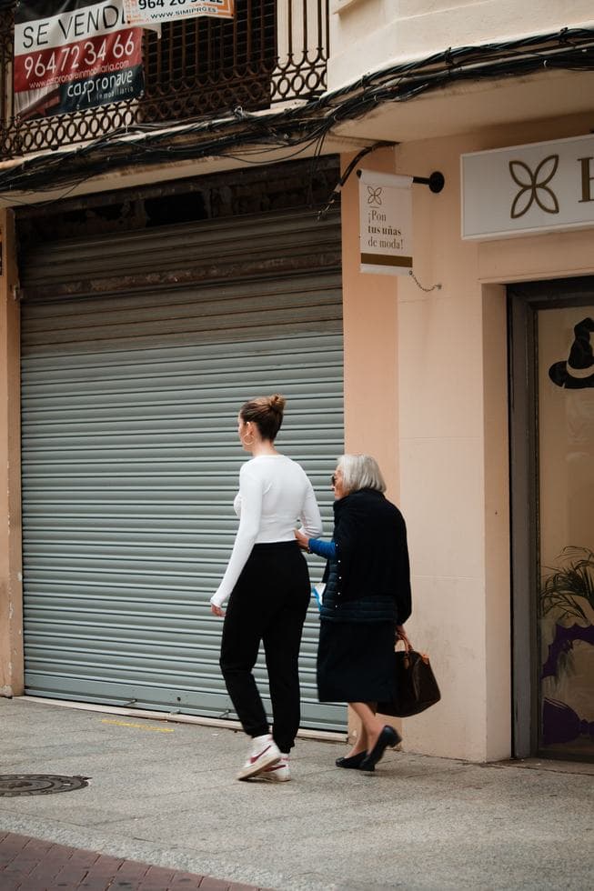 A younger woman, seen from behind, walks on a sidewalk while an older woman beside her rests a hand on her arm, past a shop with a for-sale banner.