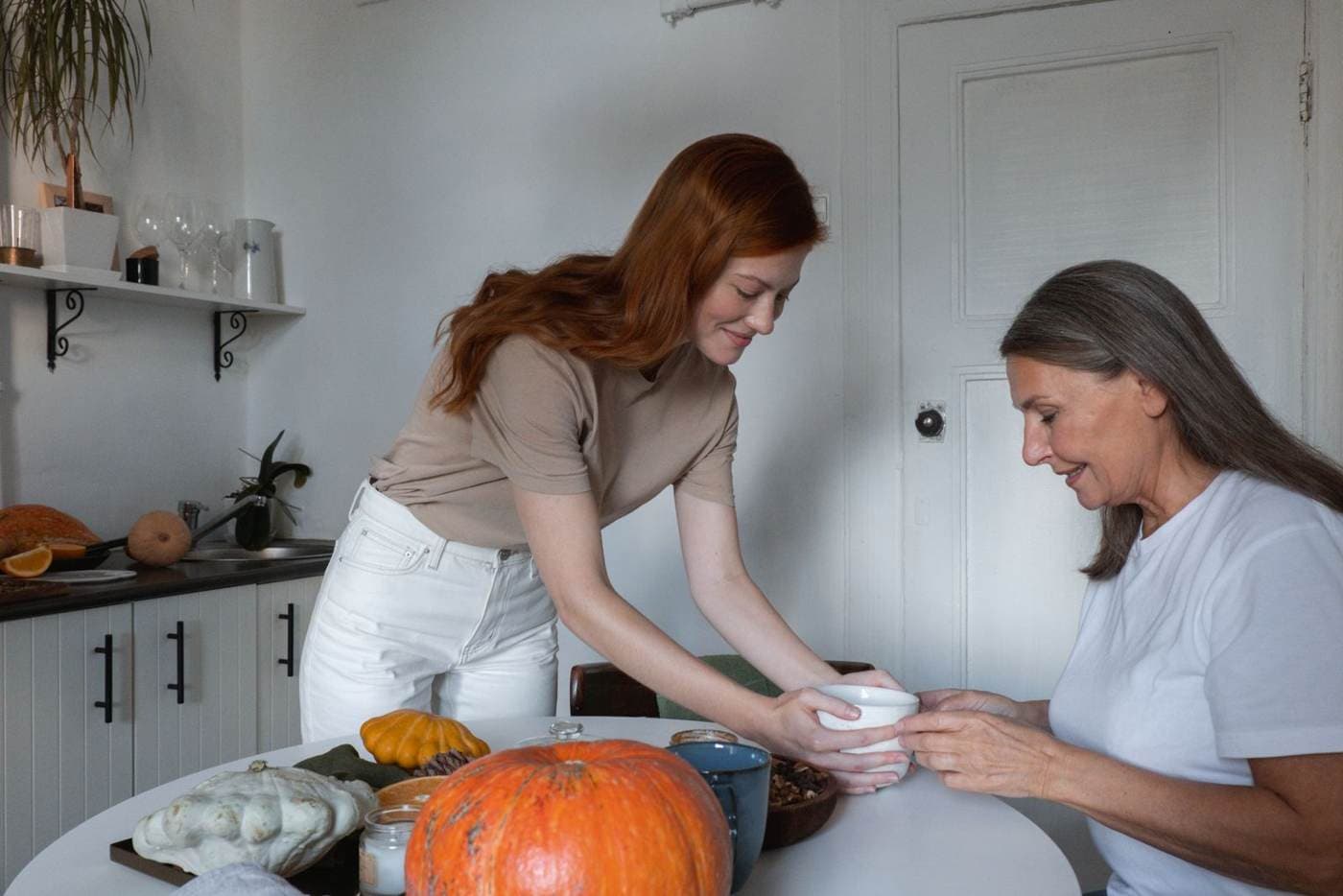 An adult daughter stands and hands a cup across a table to her seated older mother. Small pumpkins sit on the table.