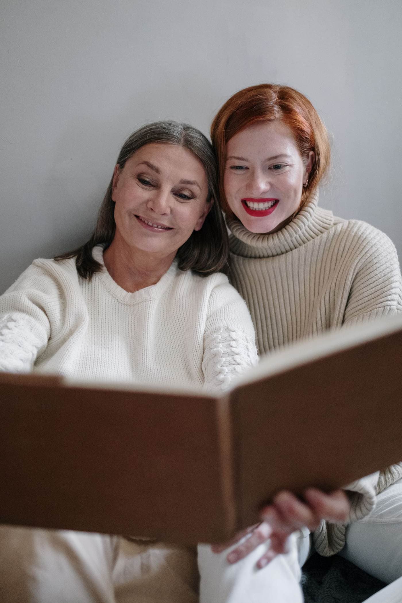 An older woman with shoulder-length gray-brown hair and a younger woman with red hair sit side by side indoors, smiling as they look at an open book they hold together.