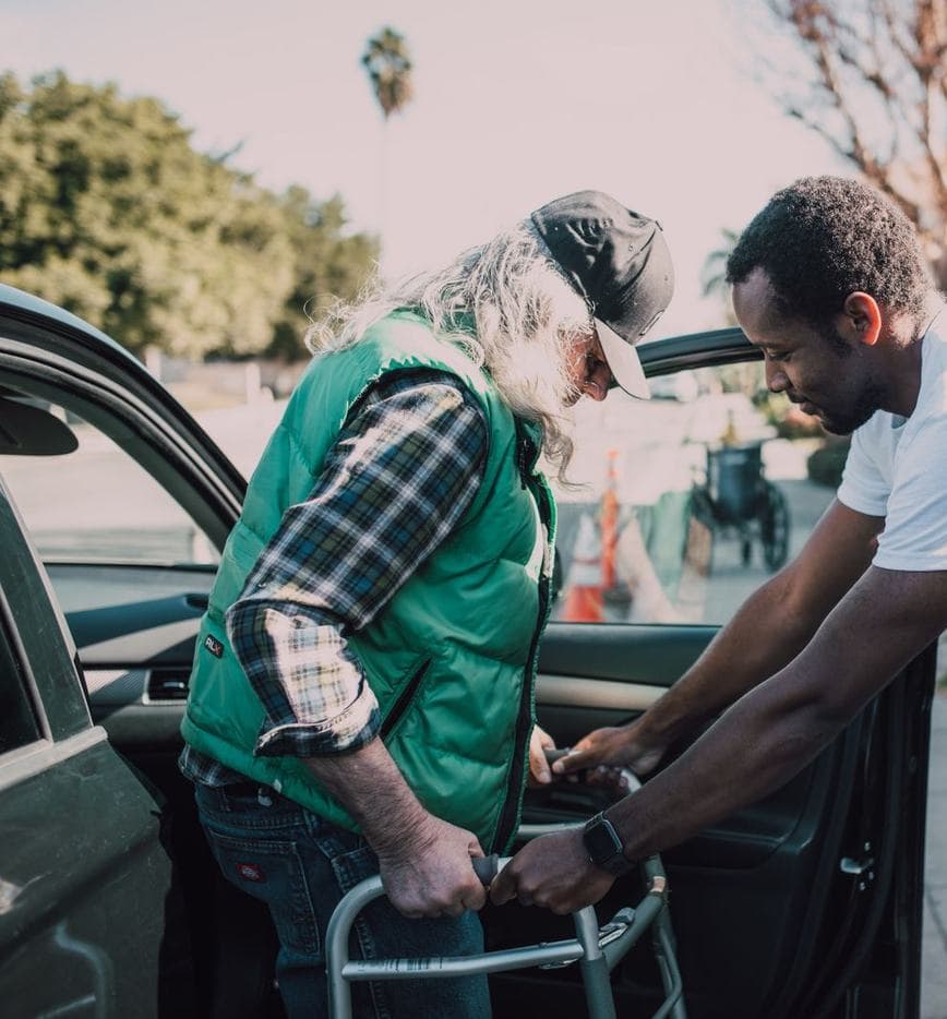 An older man looks down and holds a walker beside an open car door while a younger man grips the walker frame; a wheelchair is in the background.