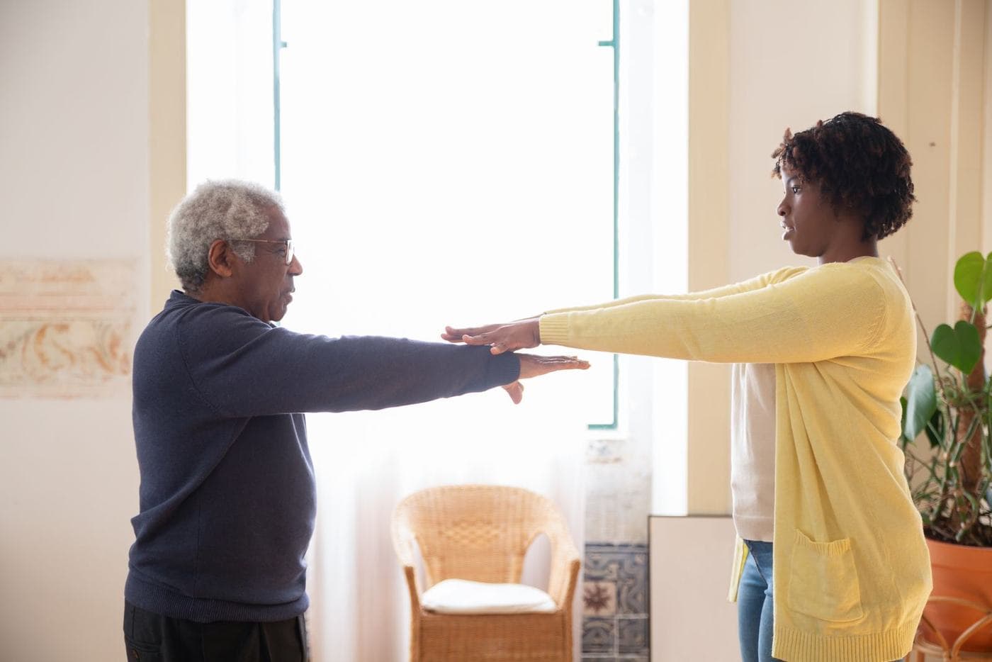 An older man and a younger woman stand in profile indoors, arms extended, his palms up and her palms just above his.