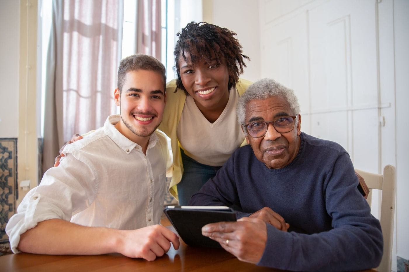 A young man and a young woman smile at the camera beside an older man at a wooden table; she rests a hand on each man's shoulder, and he holds a tablet.