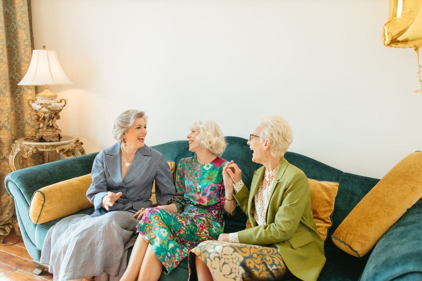 Three older women laugh together on a teal sofa, turned toward one another in a living room.