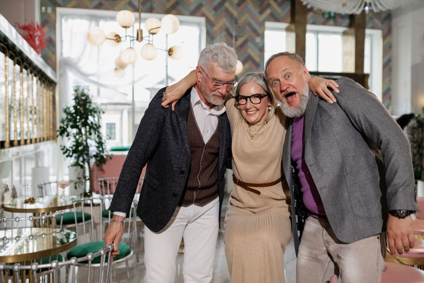 Three people in a restaurant stand close with their arms around one another; a woman and a man face the camera while an older man looks down.